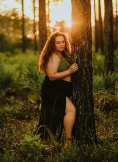 a woman leaning against a tree in the woods at sunset with her legs spread out