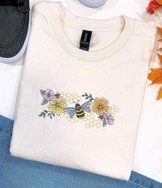 Introducing our Bee's And Flowers Embroidered T-Shirt! This playful tee features a charming design of buzzing bees and blooming flowers. Made with high-quality materials, this shirt is both stylish and comfortable. Perfect for adding some whimsy to any wardrobe. Spring T-shirt With Multicolor Embroidered Text, Cute Embroidered T-shirt For Spring, Multicolor Embroidered Text T-shirt For Spring, Spring Crew Neck T-shirt With Machine Embroidery, Cute Floral Embroidery Crew Neck T-shirt, Spring Crew Neck T-shirt With Butterfly Embroidery, Spring Butterfly Embroidered Crew Neck T-shirt, Cute Crew Neck T-shirt With Floral Embroidery, Spring Custom Embroidery Crew Neck T-shirt