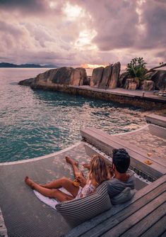 a man and woman sitting on a wooden bench next to the ocean at sunset or sunrise