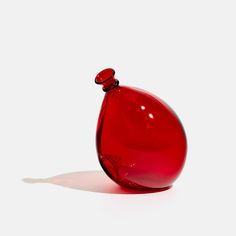 a red glass vase sitting on top of a white table next to a shadowless wall