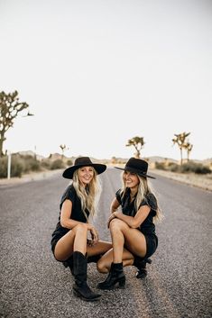 two women sitting on the side of an empty road wearing cowboy hats and black dresses