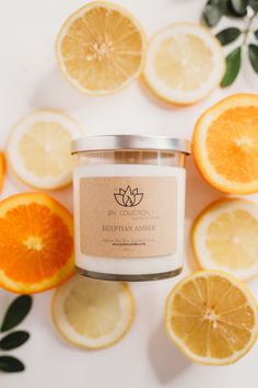 an orange and lemon scented candle surrounded by sliced oranges on a white surface with green leaves