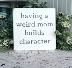 a sign that says having a weird mom build's character next to some plants