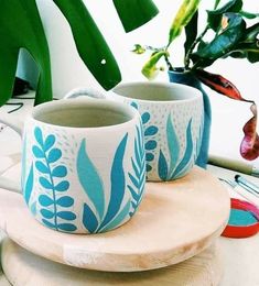 two coffee mugs sitting on top of a wooden coaster next to a potted plant