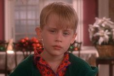 a young boy with blonde hair wearing a green sweater and looking at the camera while standing in front of a dining room table