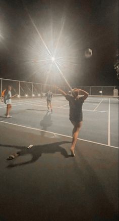 three people are playing tennis at night on an asphalt court with lights in the background