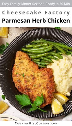 a black plate topped with chicken, mashed potatoes and green beans next to lemon wedges