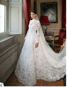 a woman in a white wedding gown standing by a window with her veil pulled back