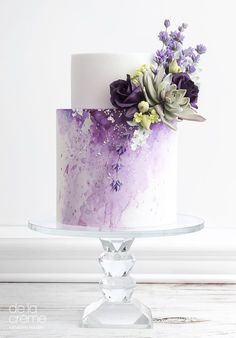 a white and purple cake with succulents on the top is sitting on a glass stand