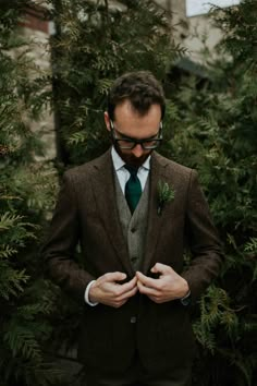 a man wearing a suit and tie standing in front of some trees with his hands on his chest