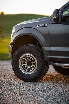 the front end of a gray truck parked on top of a gravel road next to a green field