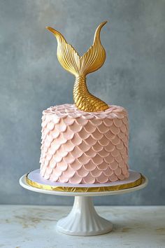 a pink and gold cake with a mermaid tail decoration on top, sitting on a white pedestal