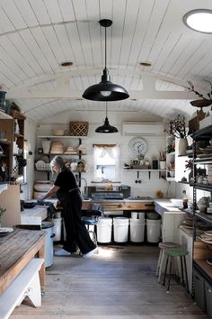 a woman standing in a kitchen next to a wooden table and shelves filled with pots