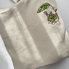 A beige Korok Hoodie by petimint, featuring an embroidered design of a small green leaf character hanging from a larger green leaf, is ideal for gaming enthusiasts. Positioned on a white surface, the stylish hoodie is bathed in natural light that casts subtle shadows across it. Green Casual Hoodie With Custom Embroidery, Casual Green Hoodie With Custom Embroidery, Cotton Hoodie With Embroidered Patch For Streetwear, Green Cotton Hoodie With Embroidered Graphics, Winter Cotton Hoodie With Embroidered Patch, Casual Hoodie With Embroidered Patch, Winter Cotton Tops With Embroidered Patch, Winter Cotton Top With Embroidered Patch, Casual Hoodie With Embroidered Patch For Winter