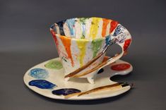 a cup and saucer on a plate with paintbrushes next to it in front of a gray background