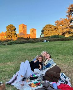 two women are sitting on a blanket in the middle of a field with trees and buildings behind them