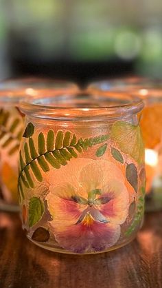 three glass jars with flowers painted on them sitting on a wooden table next to candles