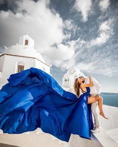 a man and woman kissing on the roof of a building with a blue dress blowing in the wind