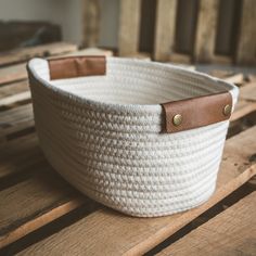 a white basket with leather handles sitting on top of a wooden table next to some wood planks