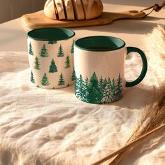 two green and white coffee mugs sitting on top of a bed next to a loaf of bread