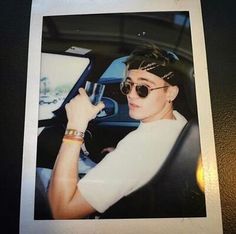 a woman wearing sunglasses sitting in a car with her hand up to the steering wheel