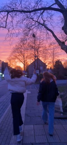 two girls walking down the sidewalk at sunset with their arms in the air and one girl holding up her hand