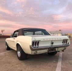 an old white mustang parked in a parking lot with the sun going down behind it