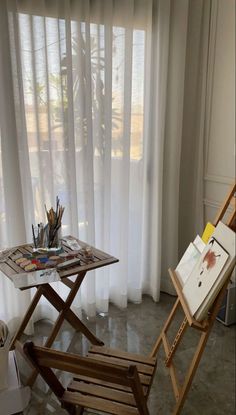 an easel and painting supplies sit on a table in front of a window with white curtains