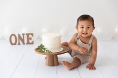 a baby sitting in front of a cake on a wooden stand with the number one sign behind it