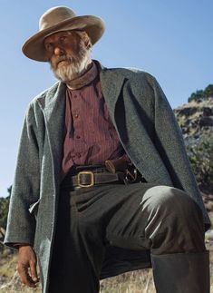 an old man in a hat and coat is sitting on a rock with his legs crossed