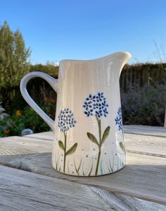 a white pitcher with blue flowers painted on the side sitting on a wooden table outside