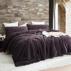 a bed with a blanket and pillows on it in front of a brick wall next to a window