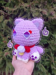 a hand holding a purple crocheted teddy bear in front of a green tree