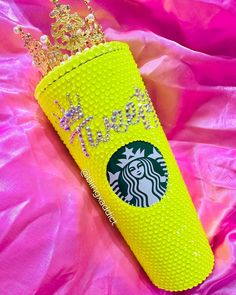 a yellow starbucks cup with a gold crown on it's head sitting on pink fabric