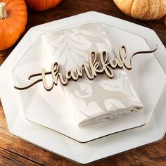 a white plate topped with a napkin covered in the word thanksgiving next to pumpkins