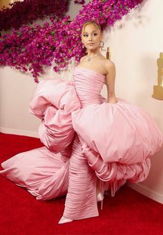 a woman in a pink gown standing on a red carpet with purple flowers behind her