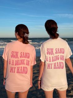 two women standing on the beach wearing matching shirts that say sun sand and a drink in my hand