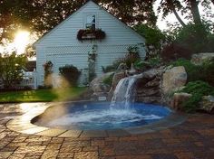 an outdoor hot tub with a waterfall in the middle and landscaping around it, near a house