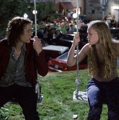 a man and woman sitting on swings in front of some parked cars, one holding onto the strings