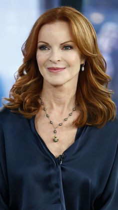 a woman with red hair wearing a blue shirt and smiling at the camera while standing in front of a tv screen