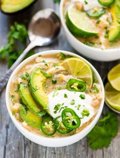 two white bowls filled with soup and topped with sour cream, avocado slices, cilantro, green peppers, and limes