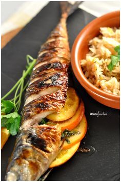 a fish and rice dish on a black plate with a bowl full of vegetables next to it