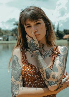 a woman with tattoos on her body posing for a photo near the water and holding her hand up to her face