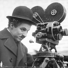 a man wearing a suit and hat looking at an old movie projector on a tripod