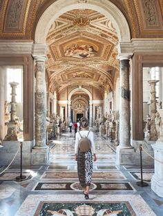 a woman is walking down the hall in an ornate building with columns and paintings on the walls