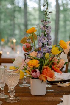 an arrangement of colorful flowers in a white vase on a wooden table with wine glasses