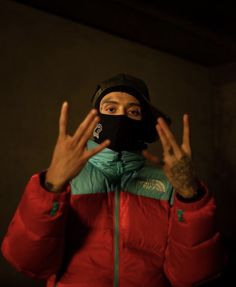 a man in a red jacket and black hat covering his face with both hands while making the vulcan sign