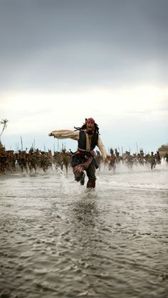 a man in pirate garb running through the water with a surfboard on his back