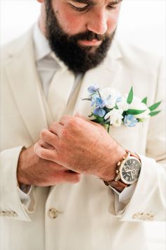 a man wearing a white suit and blue flower boutonniere is looking down at his watch