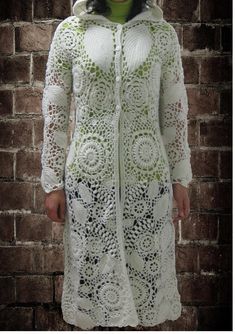 a woman standing in front of a brick wall wearing a white crocheted coat